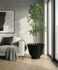 A modern living room features a light gray couch with a dark gray pillow and a beige pillow, draped with a gray knit blanket. Beside the couch stands a tall, elegant black planter pot filled with a lush green tree, positioned near a large window that lets in natural light. A framed abstract artwork hangs on the concrete-textured wall behind the couch. The room is finished with light wood flooring.