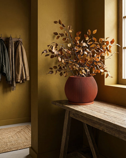 A terracotta planter pot sits on a wooden table, filled with dried leaves. Coats hang on a wall in the background.