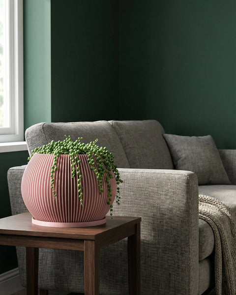 A pink planter pot with a string of pearls plant sits on a wooden table next to a gray couch in a room with dark green walls.