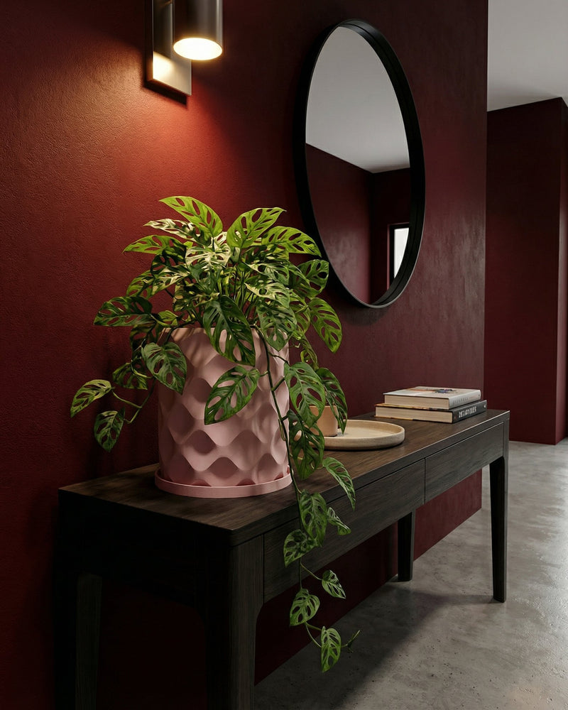 A pink planter pot with a green plant sits on a dark wood table against a red wall with a mirror and light fixture.