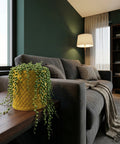A yellow planter pot with a string of pearls plant sits on a wooden table next to a gray sofa.