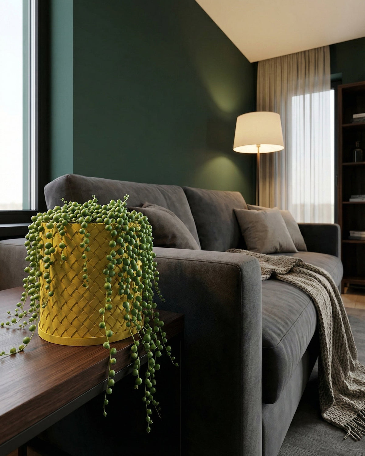A yellow planter pot with a string of pearls plant sits on a wooden table next to a gray sofa.