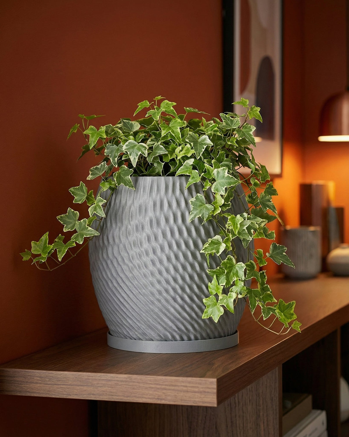 A gray planter pot with a green ivy plant sits on a wooden shelf against a rust-colored wall.