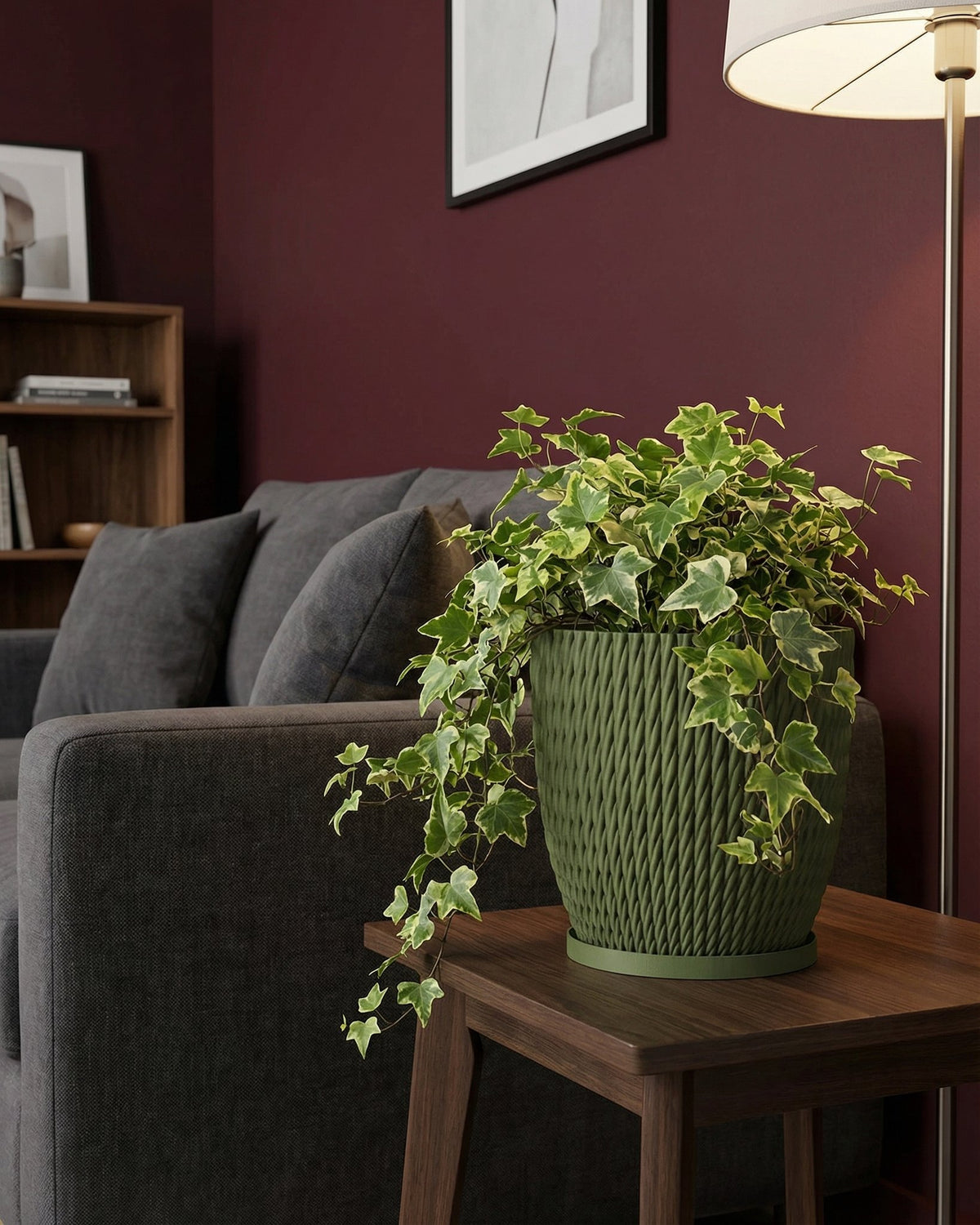 A green ivy plant in a green woven planter pot sits on a dark wood side table next to a gray couch in a living room.