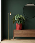 A red planter pot with a green plant sits on a wooden table next to a lamp. The pot has a textured design.