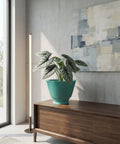 A green planter pot with a plant inside sits on top of a wooden cabinet. The cabinet is in a room with a concrete wall and a painting on the wall. A floor lamp is next to the cabinet.
