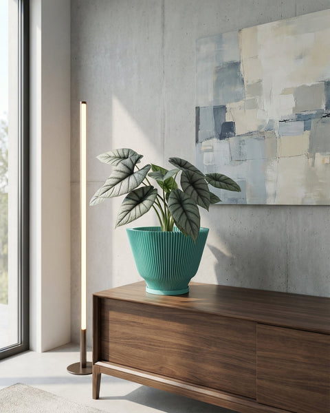 A green planter pot with a plant inside sits on top of a wooden cabinet. The cabinet is in a room with a concrete wall and a painting on the wall. A floor lamp is next to the cabinet.