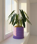 A purple planter pot with a plant inside sits on a white windowsill in front of a window.