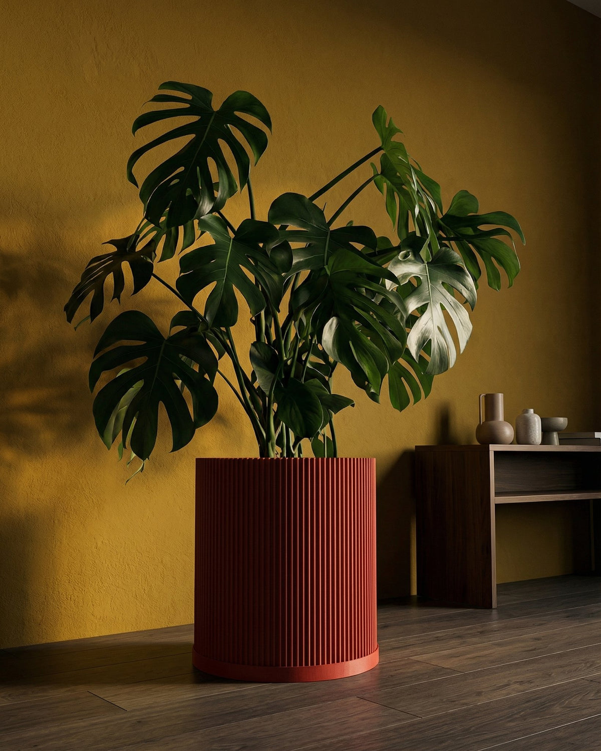 A large Monstera plant in a red, ridged planter pot sits against a yellow wall, with a wooden shelf and vases in the background.