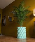 A tall green palm plant sits in a light blue planter pot with a geometric pattern. The pot is on a wooden floor in a room with yellow walls.