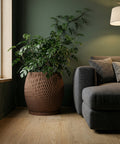 A brown textured planter pot with a green plant next to a gray couch on a wooden floor.
