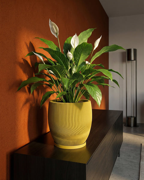A peace lily in a yellow planter pot sits on a dark wood cabinet against an orange wall. The pot features a wavy, ribbed design.