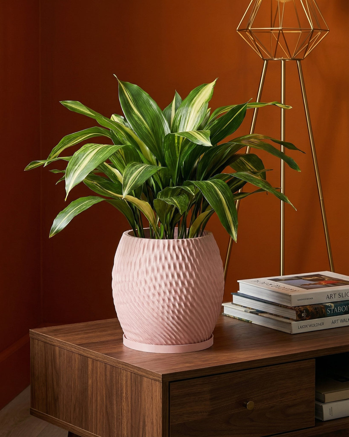 A pink planter pot with a green plant sits on a wooden table next to books and a gold lamp.