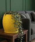 A yellow planter pot with a trailing plant sits on a wooden table next to a gray sofa. The pot has a textured, swirling design.