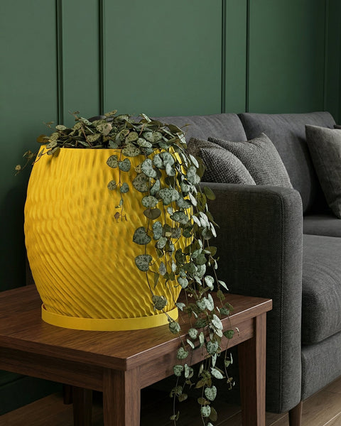 A yellow planter pot with a trailing plant sits on a wooden table next to a gray sofa. The pot has a textured, swirling design.