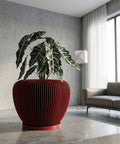 A red planter pot with a plant inside sits on a concrete surface in a modern room. The pot has a ribbed design.