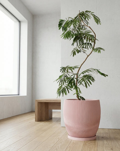A pink planter pot with a green plant inside, sitting on a light wood floor next to a white wall.