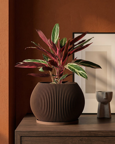 A planter pot with a plant in it sits on a wooden table next to a small sculpture. The planter pot is brown and has a unique design.