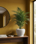A vibrant green palm plant sits in a stylish, white ribbed planter pot on a dark wood table against a mustard-colored wall.
