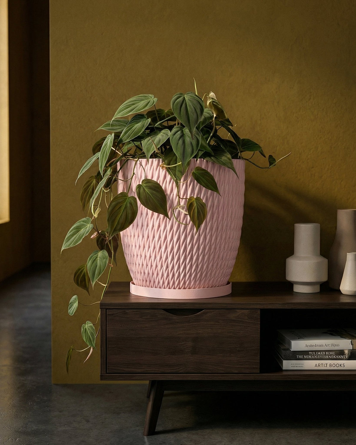 A pink planter pot with a plant inside sits on a dark wood cabinet with books and vases.