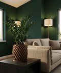 A ZZ plant in a brown planter pot sits on a dark wood table next to a beige sofa in a room with dark green walls.