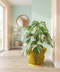 A yellow planter pot with a green plant sits on a wooden table next to a gray couch with a throw blanket.