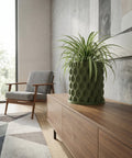 A green planter pot with a spider plant sits atop a wooden cabinet in a modern living room with a gray chair and patterned rug.