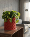 A Monstera plant in a red planter pot with a moss pole, set against a mustard-colored wall. The pot has a woven texture.