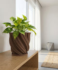A brown textured planter pot with a green plant next to a gray couch on a wooden floor.
