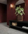 A brown geometric planter pot with a green banana plant sits on a gray shelf against a dark red wall.