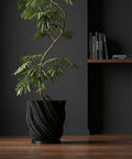 A black planter pot with a plant sits on a dark wood table. A round mirror hangs on the wall behind the plant.