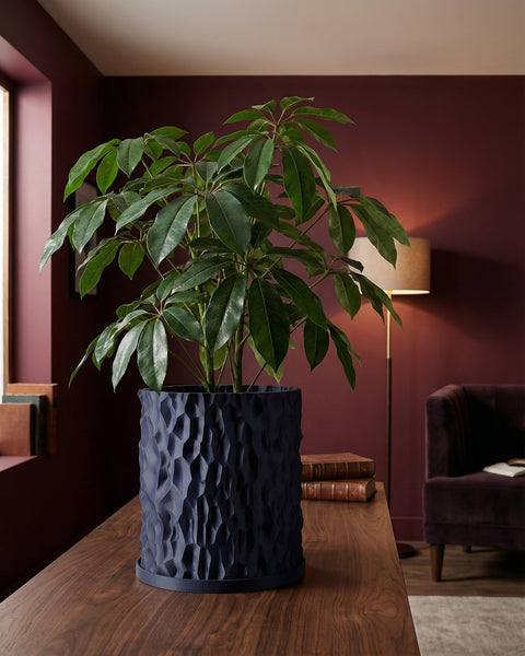 A dark blue planter pot with a textured, geometric design sits on a wooden table, holding a lush green plant. The pot is the focal point.