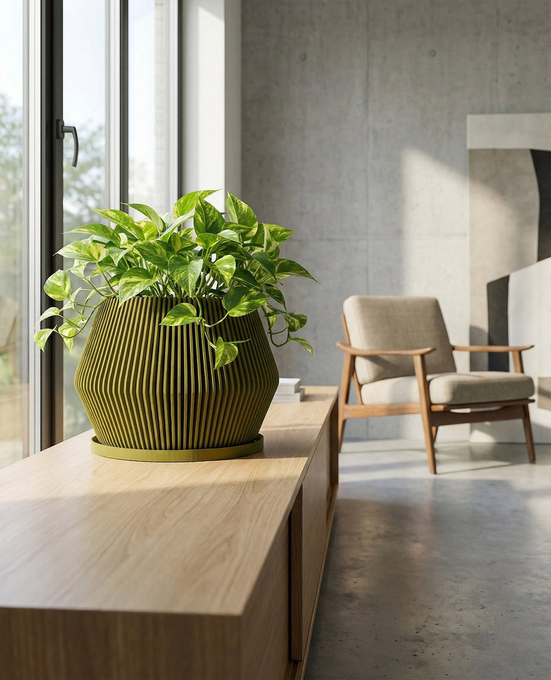 A green planter pot with a plant sits on a gray table. The planter pot has a unique design with vertical lines. In the background, there is a gray couch and a wooden coffee table. A window with sheer curtains is visible in the background.