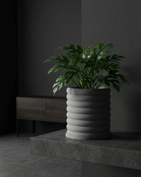 A modern gray planter pot with a ribbed design, holding a lush green monstera plant, sits on a concrete surface against a dark gray wall.