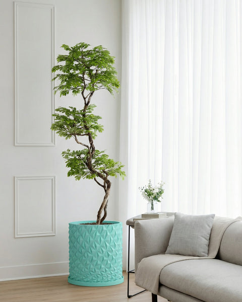 A tall, green tree in a turquoise planter pot sits next to a couch with a pillow and blanket. A small table with flowers is nearby.