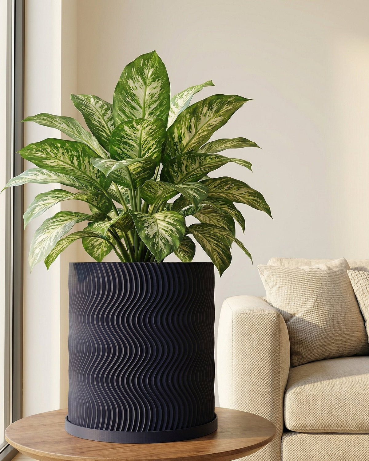 A Dieffenbachia plant in a modern, dark blue planter pot with a wavy design, sitting on a round wooden table next to a beige couch.
