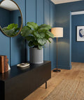 A gray planter pot with a green plant sits on a black cabinet in a blue room with a gold mirror and floor lamp.