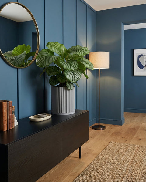 A gray planter pot with a green plant sits on a black cabinet in a blue room with a gold mirror and floor lamp.